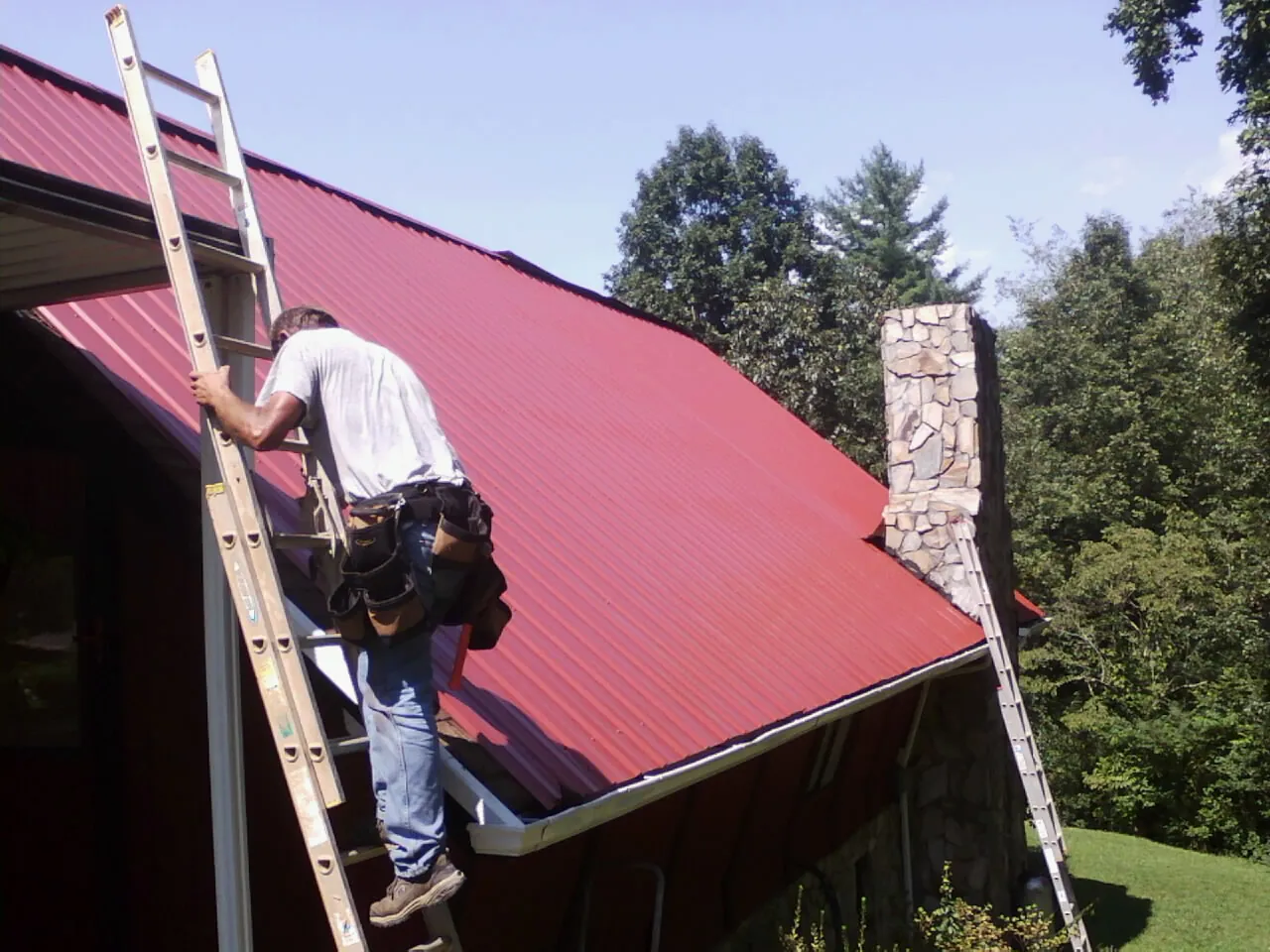 Roofer on a ladder beside a red metal roof and stone chimney — Soffit & Fascia Repair by Roofing Brothers in Ventura